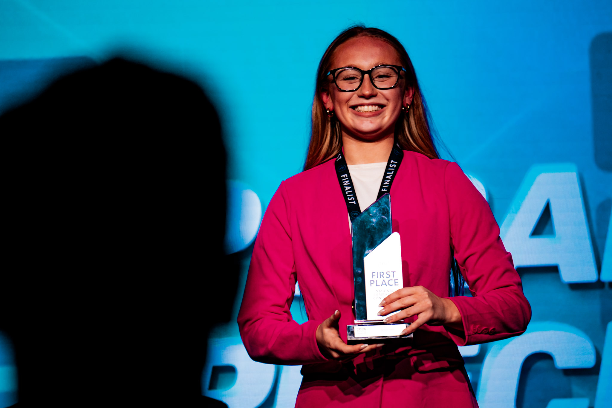 Woman in pink blazer holding First Place trophy on stage — BPA NLC